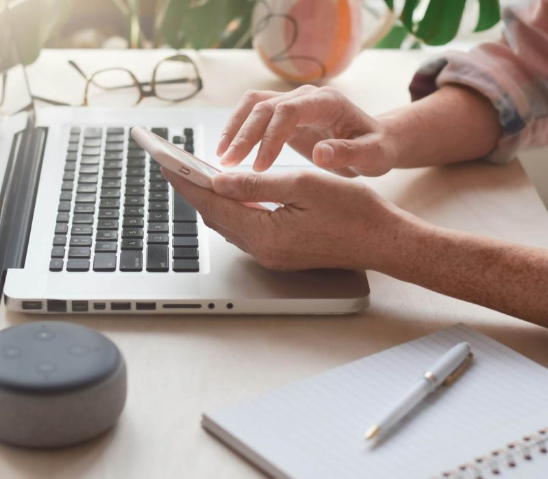 woman-sitting-at-desk-with-laptop-computer-and-virtual-assistant-smart-speaker-using-mobile-phone.jpg